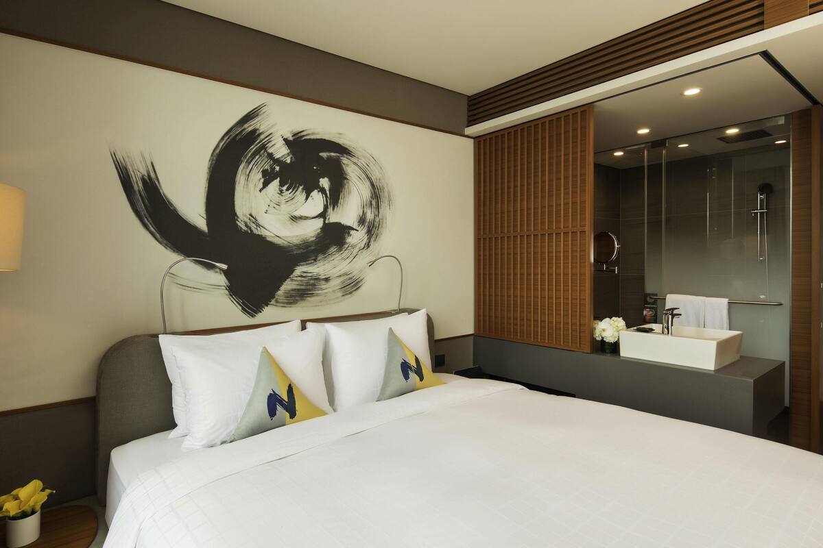 Une chambre d’hôtel comprenant un lit, un grand tableau abstrait, une salle de bain moderne avec un miroir et un lavabo, et un décor mural composé de panneaux en bois.