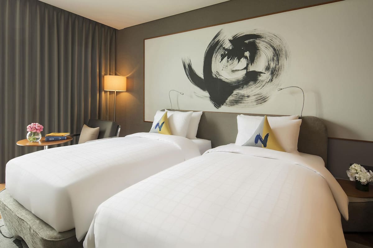 Une chambre d’hôtel avec deux lits, une grande peinture abstraite au mur, une lampe et une petite table sur laquelle est posé un vase de fleurs.