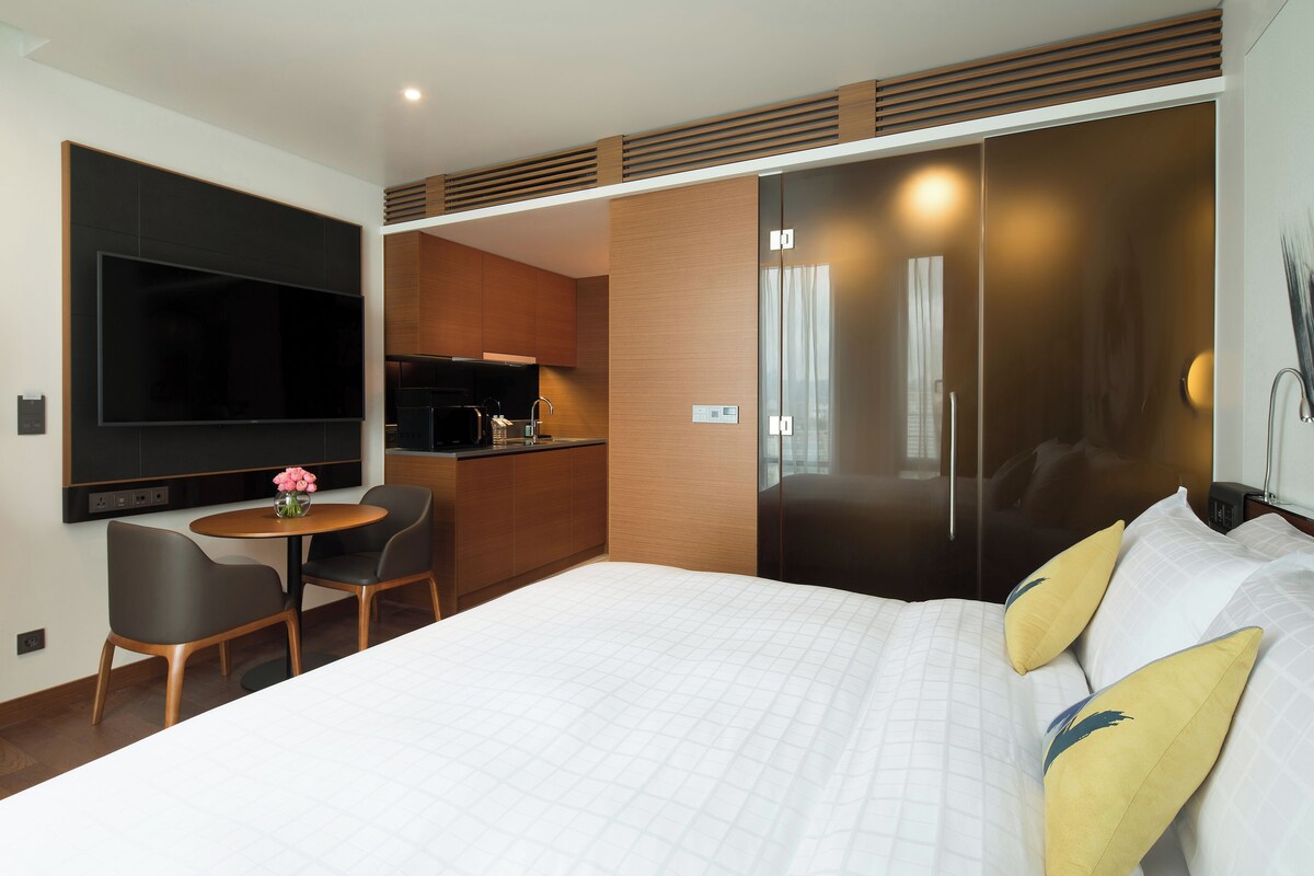 A modern hotel room with a large bed, a flat-screen TV, a small dining table, and a kitchenette.