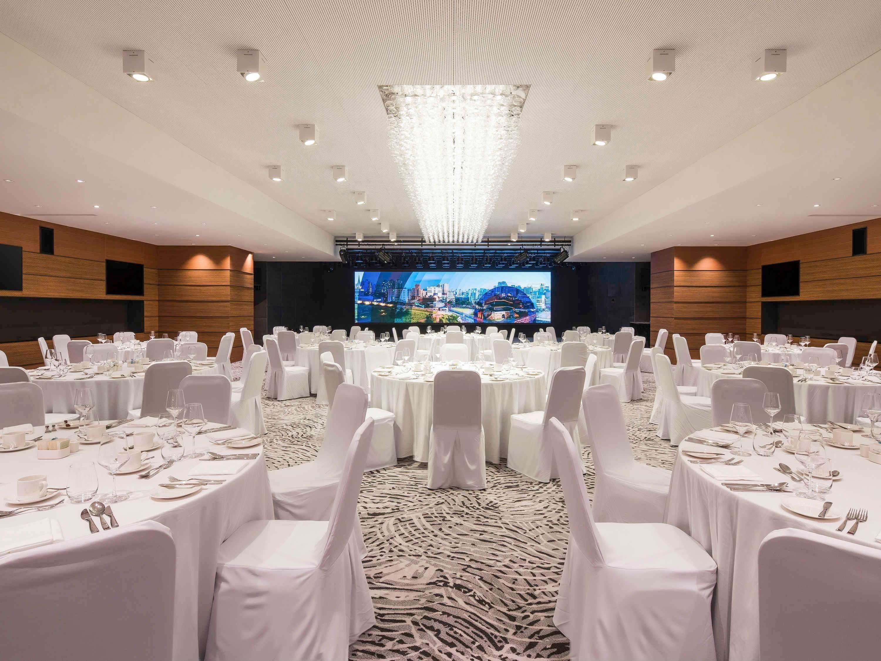 A conference room with round tables, white chairs, a large screen, and a chandelier.