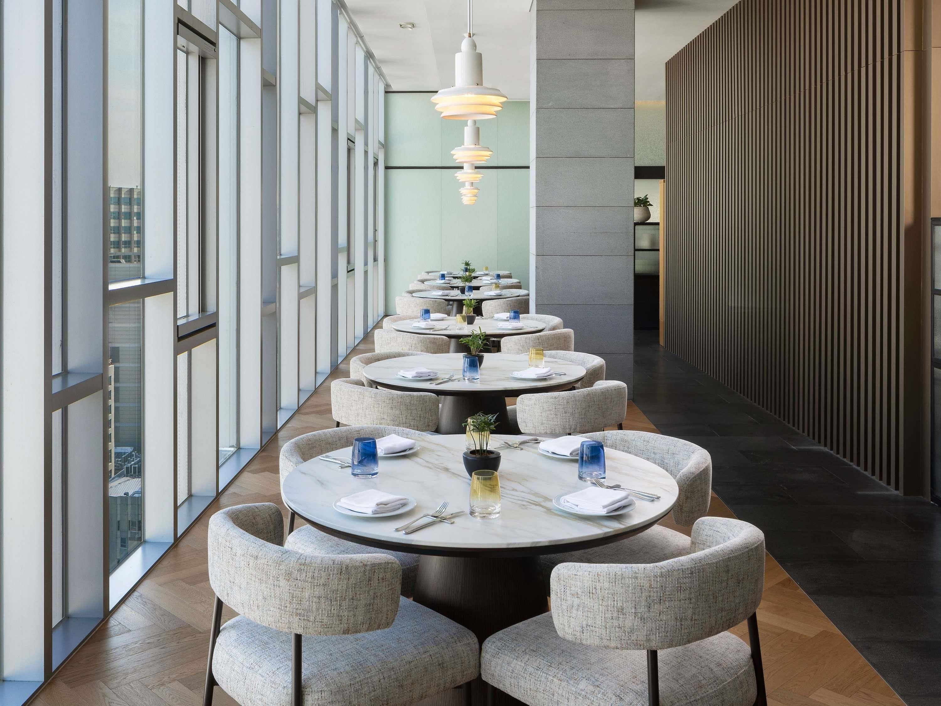 A modern dining area with round tables, grey upholstered chairs, and a long view of the city through large windows.