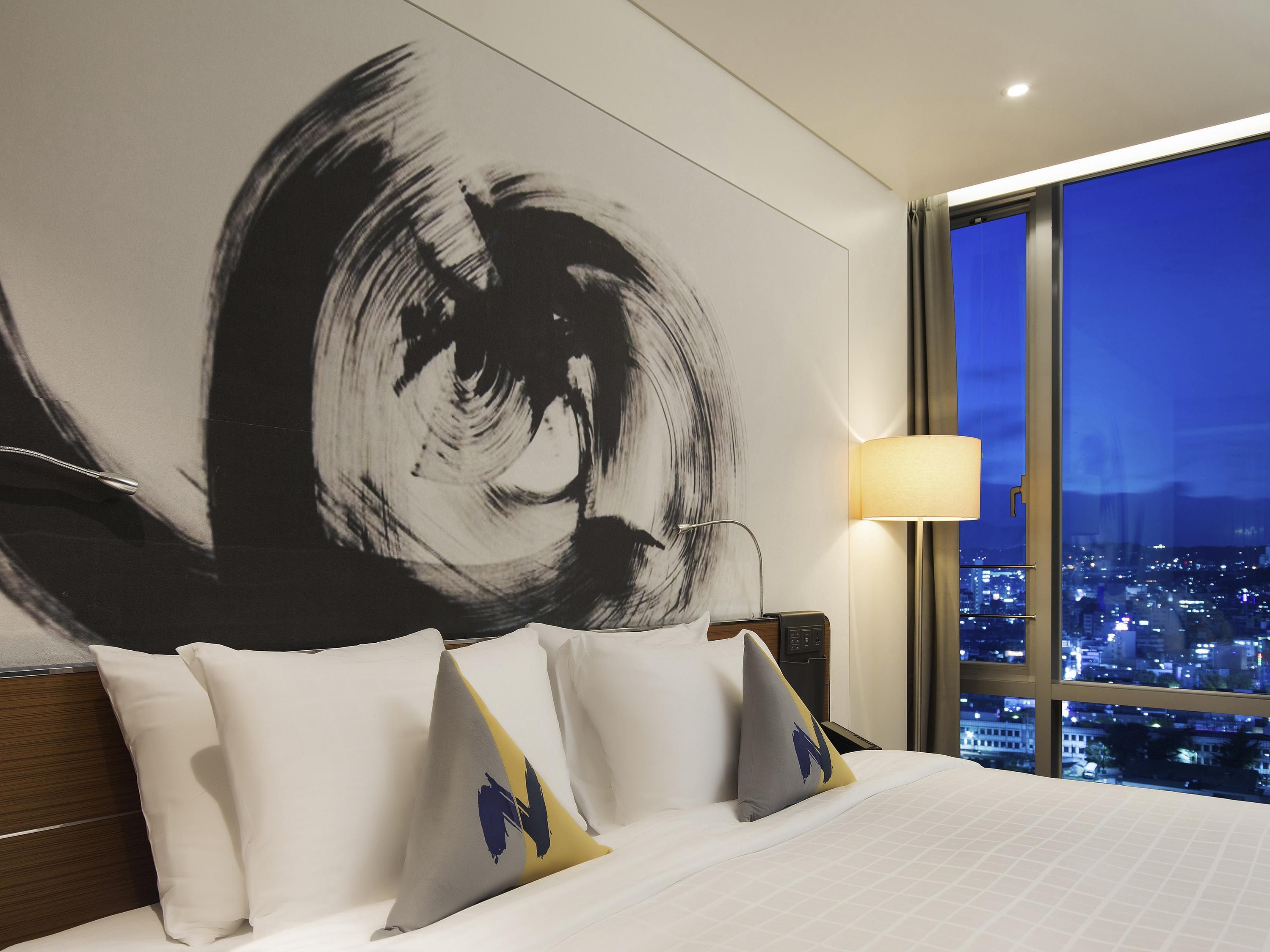 A hotel room with a large abstract painting on the wall, a bed with white bedding and decorative pillows, a bedside lamp, and a view of a cityscape at night.