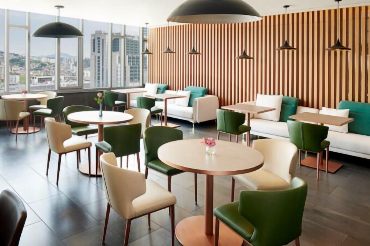 A modern lounge area with round tables and green chairs, a white sofa, and a city view through large windows.