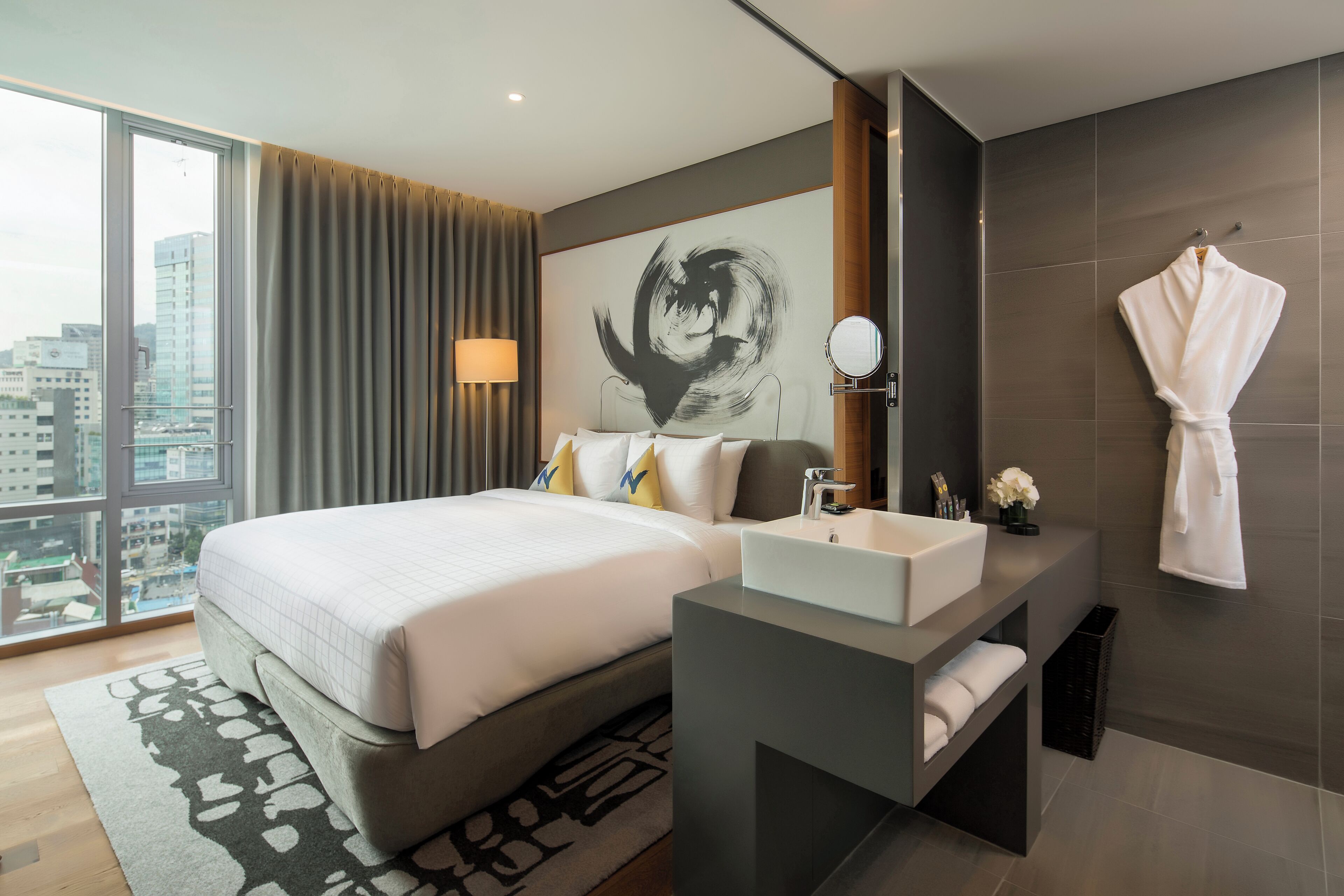A modern hotel room with a large bed, a bathroom with a sink and mirror, and a view of the city.