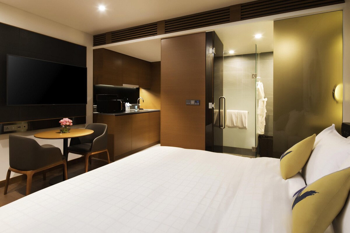 A hotel room with a bed, a desk with a chair, a TV, a kitchenette, and a shower area.