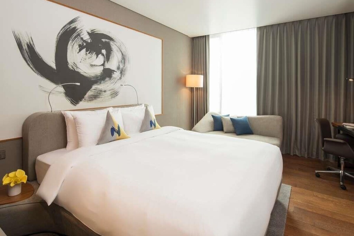 A hotel room with a large bed, a desk, a chair, a lamp, and a painting on the wall.