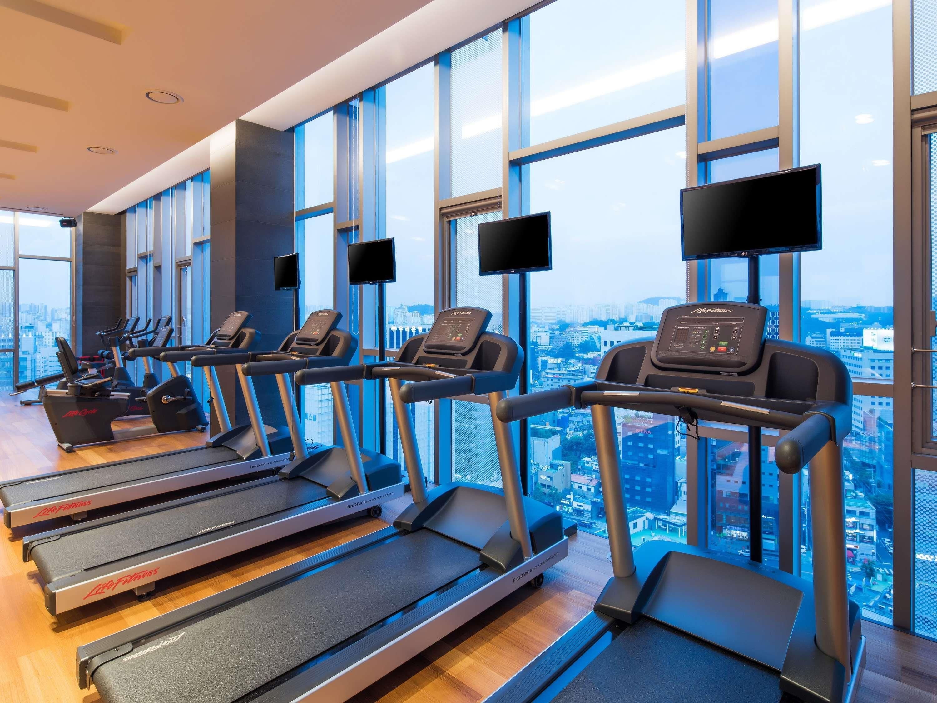 Une salle de sport équipée de tapis de course et d’appareils d’exercice, de grandes fenêtres offrant une vue sur la ville et d’un aménagement intérieur moderne.
