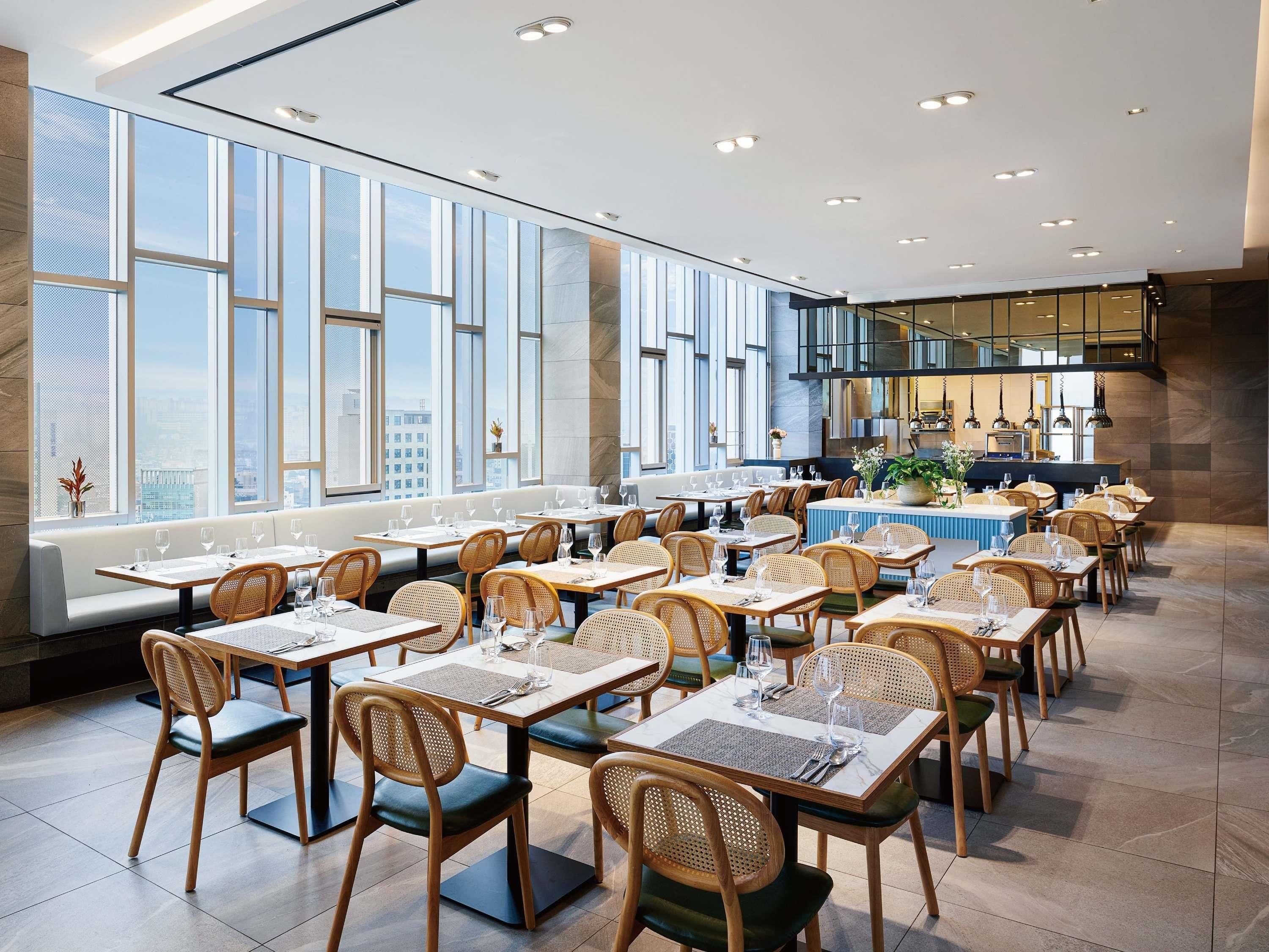 A modern restaurant with high ceilings, large windows, and a view of the cityscape. Tables are set with white tablecloths and wooden chairs.