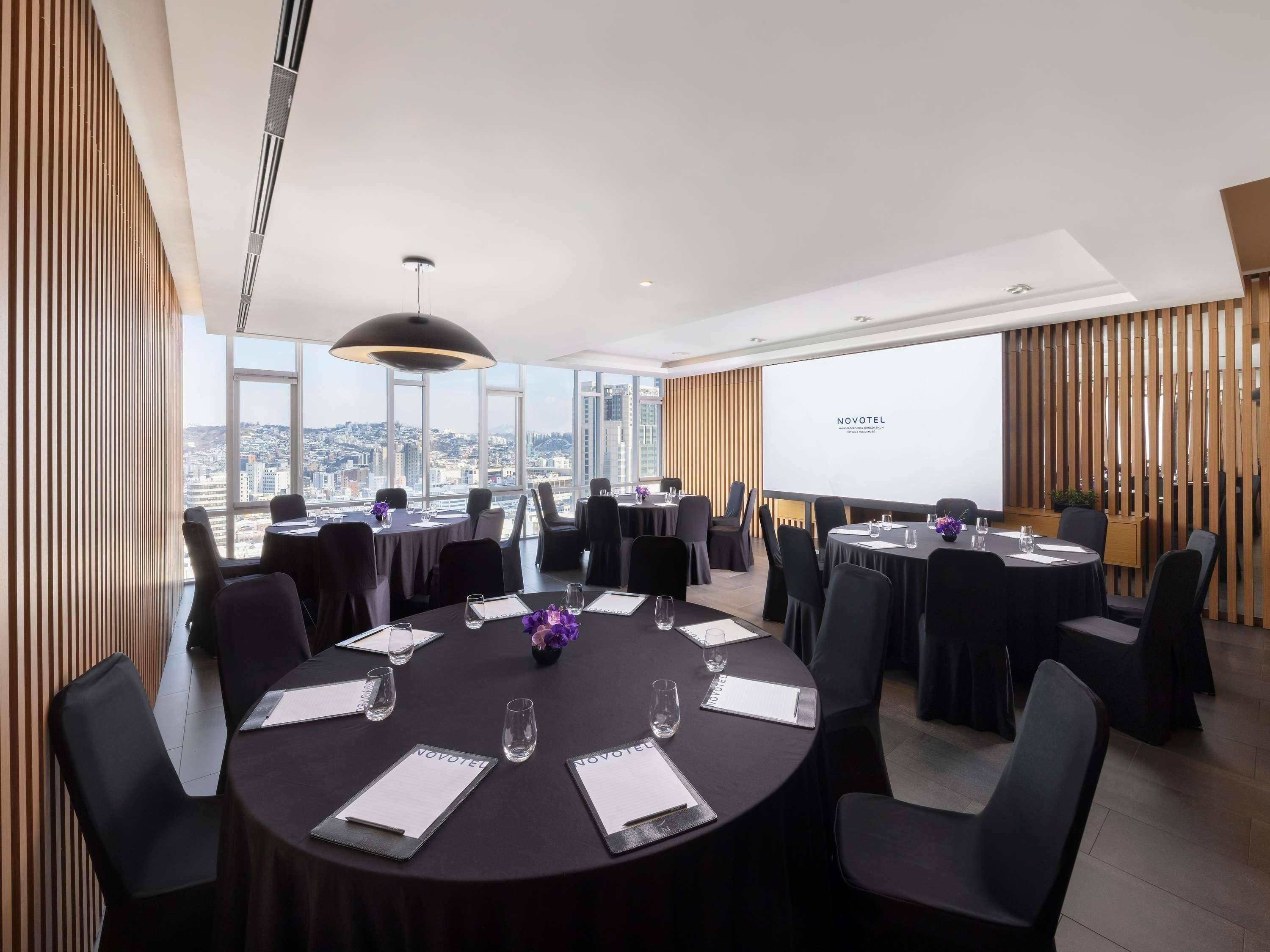 Un salón de conferencias moderno con mesas redondas, sillas negras y vistas a la ciudad.