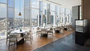A modern hotel room with large windows offering a city view, a dining area with round tables and chairs, and a kitchenette. - Novotel Ambassador Seoul Dongdaemun Hotels & Residences (Seoul)