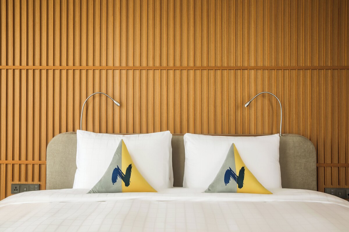 A hotel room with a wooden panel wall, a bed with white bedding, and two pillows with a yellow and blue design.