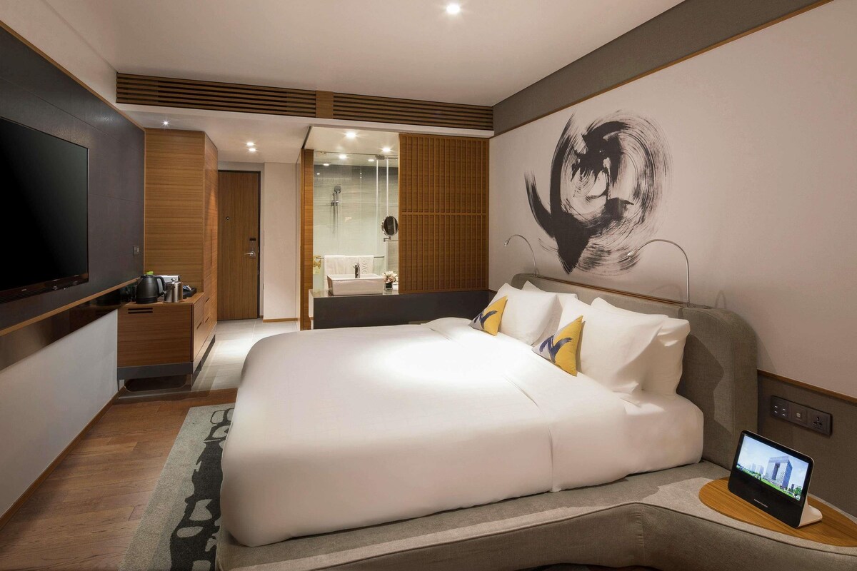 A modern hotel room with a large bed, a TV, a bathroom with a shower, and a wall art piece.