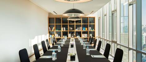 Un salón de conferencias con una mesa larga, sillas, botellas de agua y una estantería.