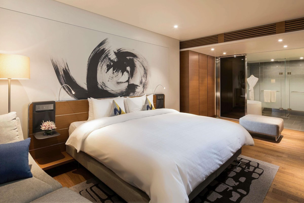 Une chambre d’hôtel moderne avec un grand lit, une table de chevet avec une lampe, une œuvre d’art murale et une douche séparée par des parois en verre.