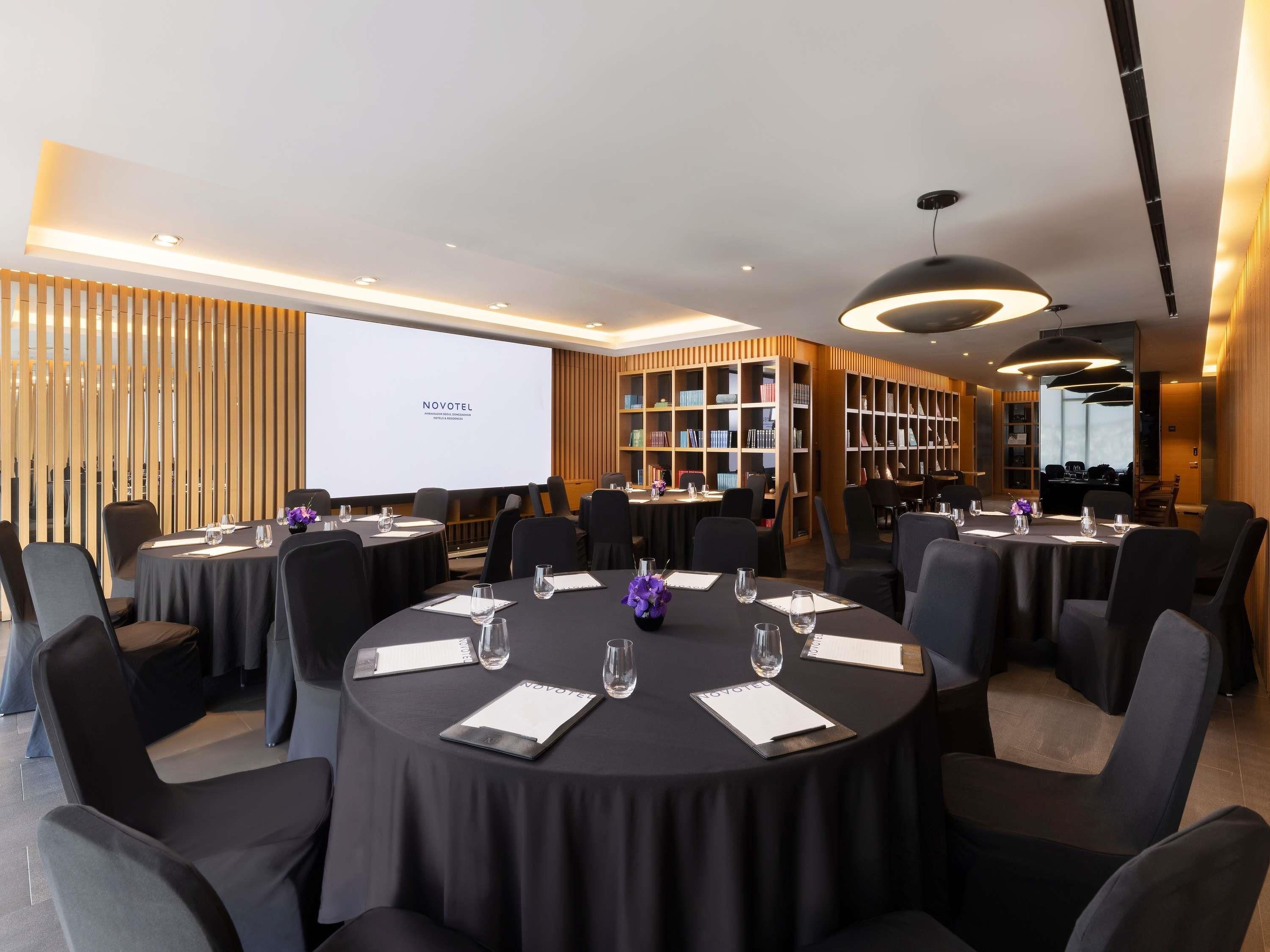 A conference room with round tables, chairs, a projector screen, and a bookshelf.