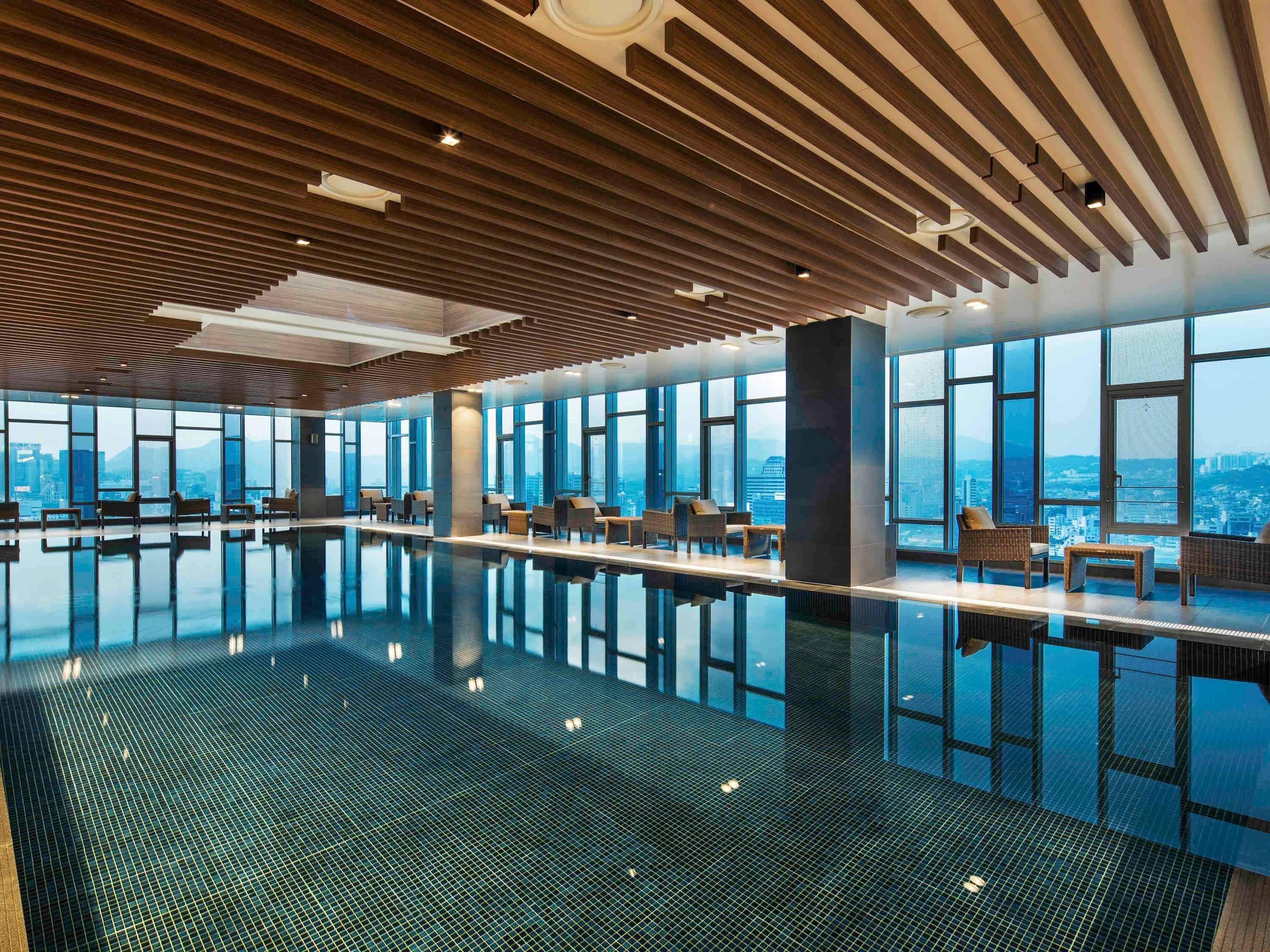 A modern indoor swimming pool with a glass floor, large windows, and seating areas.
