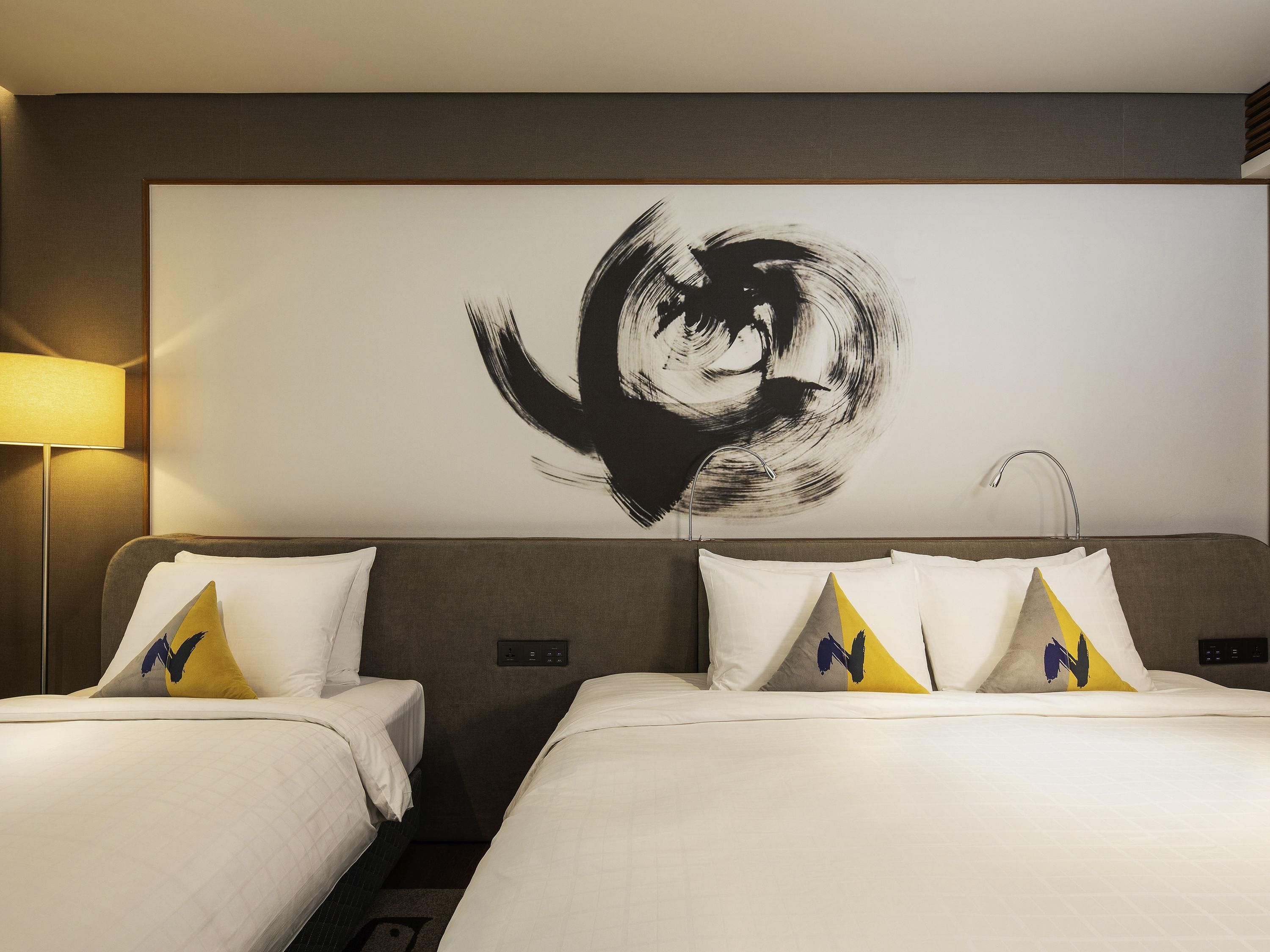 Habitación de hotel con dos camas, un cuadro abstracto grande en la pared y una lámpara a la izquierda.
