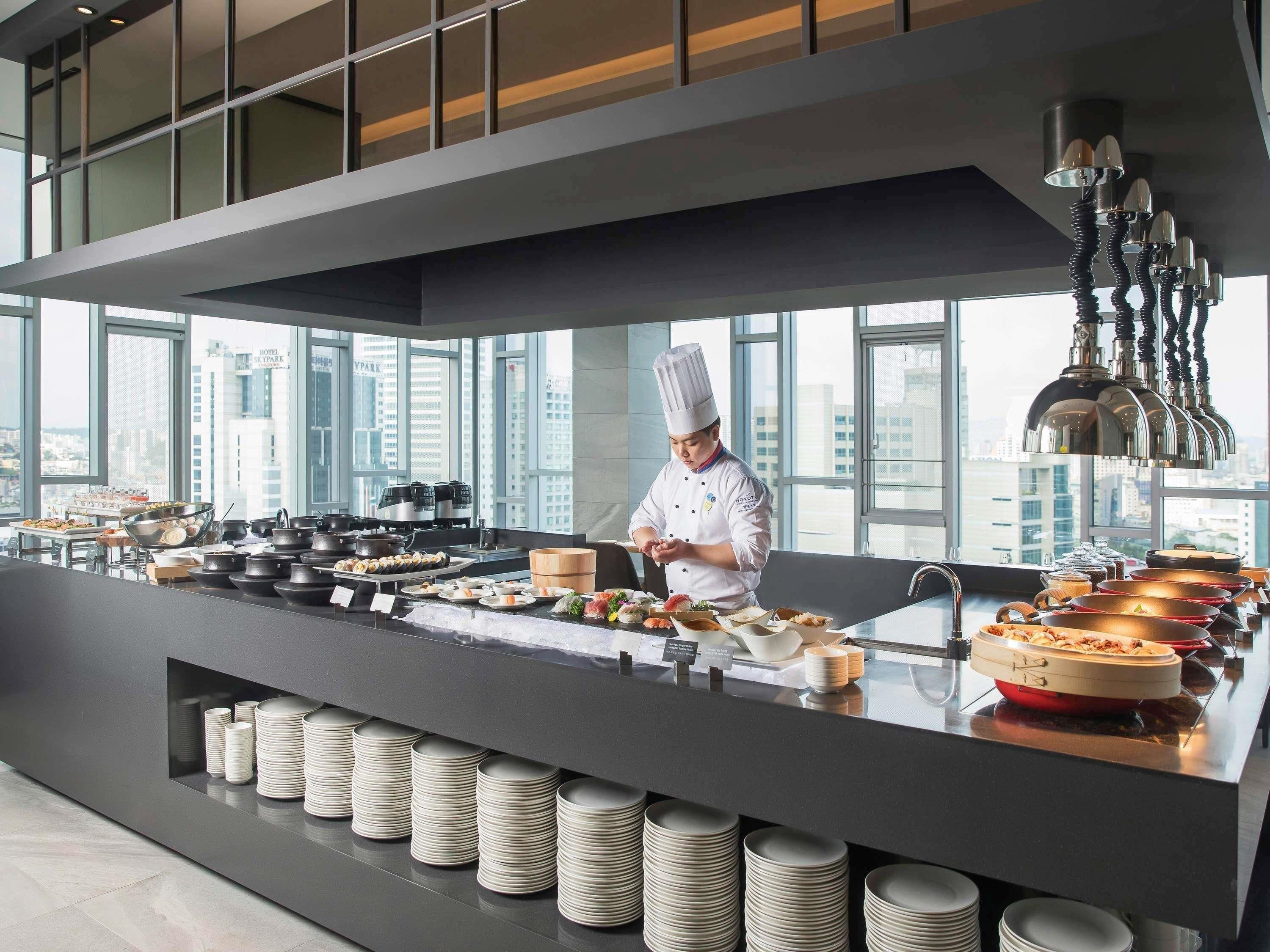 Une cuisine moderne où un chef prépare des plats, avec de grandes fenêtres et une vue sur la ville.