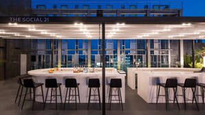 An outdoor bar area with a white counter, black bar stools, and a view of the cityscape at dusk. - Novotel Ambassador Seoul Dongdaemun Hotels & Residences (Seoul)