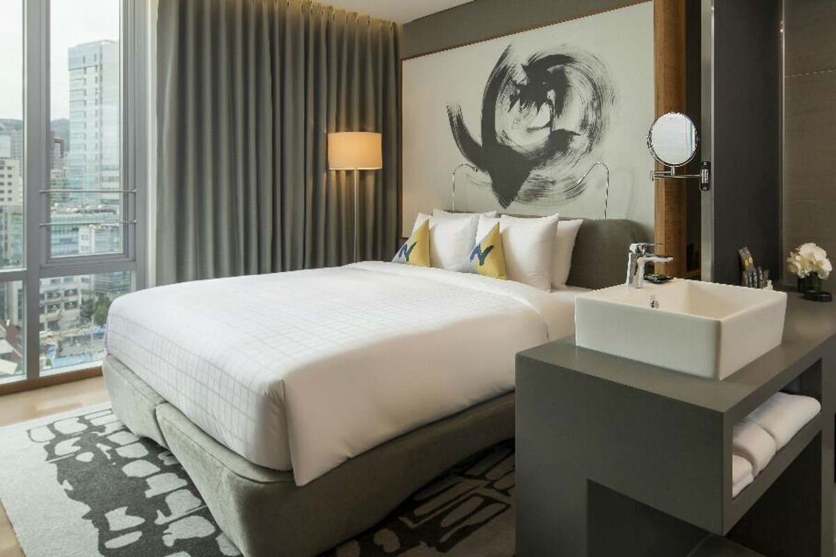 A modern hotel room with a large bed, a bedside table with a mirror, a built-in sink, and a robe hanging on the wall.
