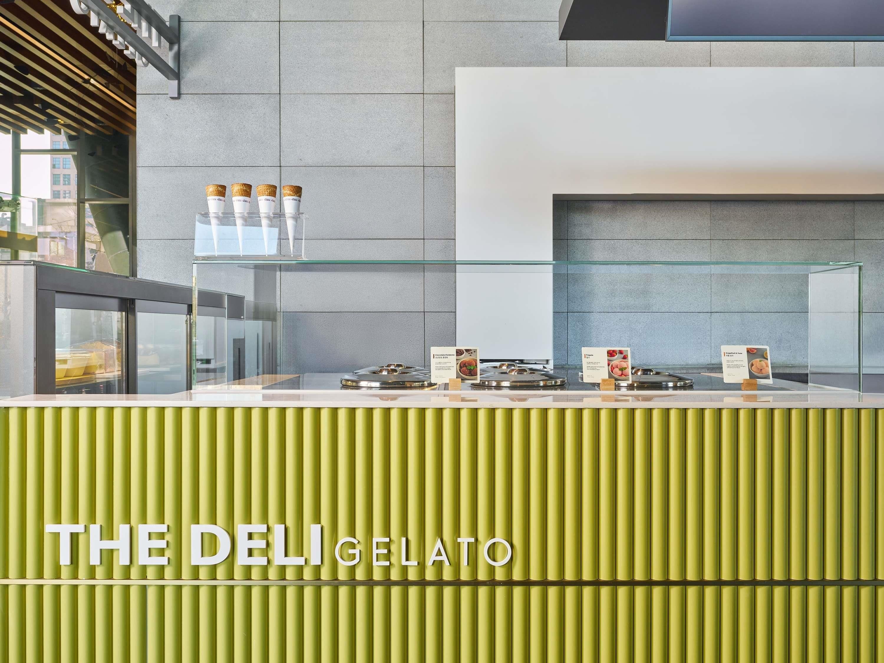 A modern deli counter with a green facade, glass display cases, and a sign reading "THE DELI GELATO".