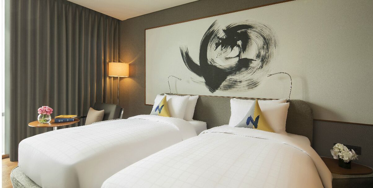 Habitación de hotel con dos camas, un cuadro abstracto grande en la pared, una lámpara y una mesita con un florero.