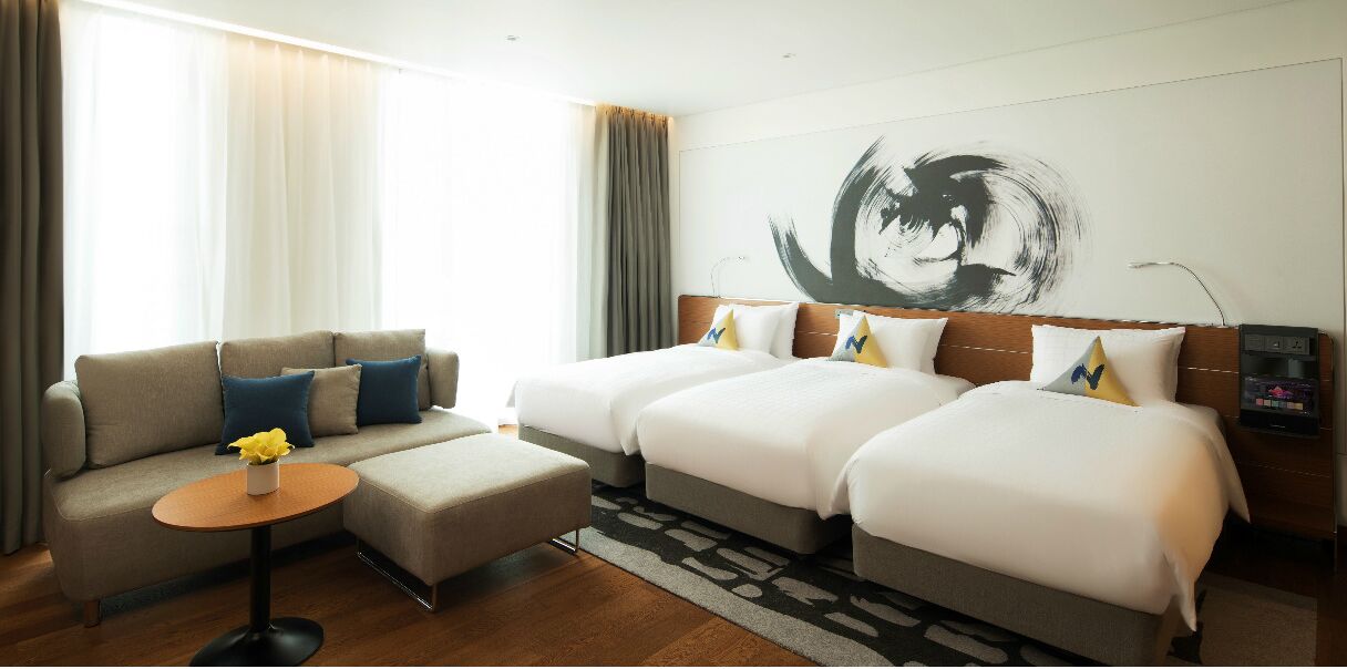 A hotel room with two beds, a sofa, a small table with a vase, and a large abstract painting on the wall.