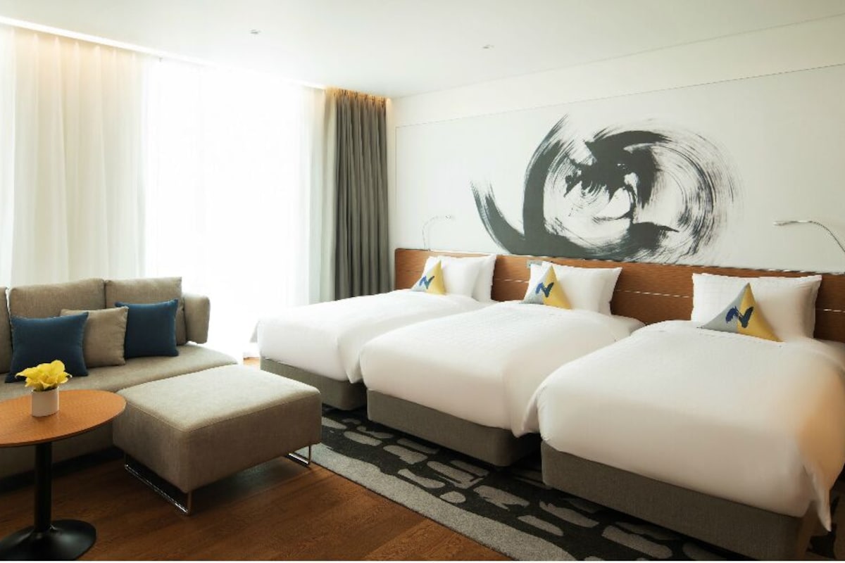 A hotel room with two beds, a sofa, a small table with a vase, and a large abstract painting on the wall.