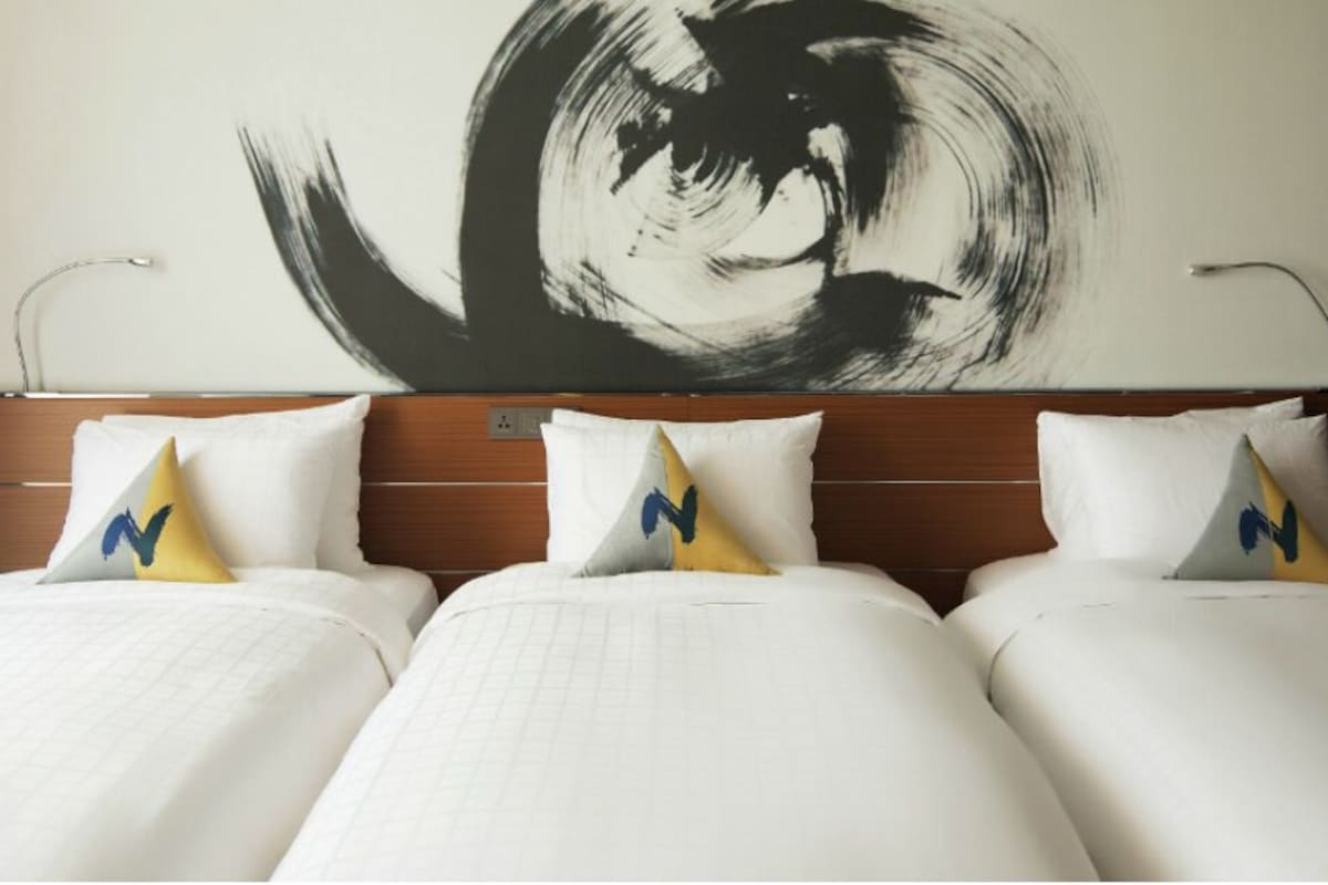 A hotel room with a large bed, three pillows, a wall art piece, and a bedside table with a lamp.