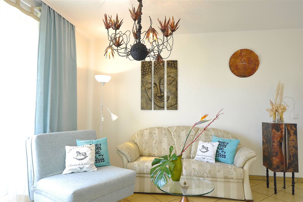 Uma sala de estar com sofá, mesa de centro, lustre, relógio e um vaso com plantas.