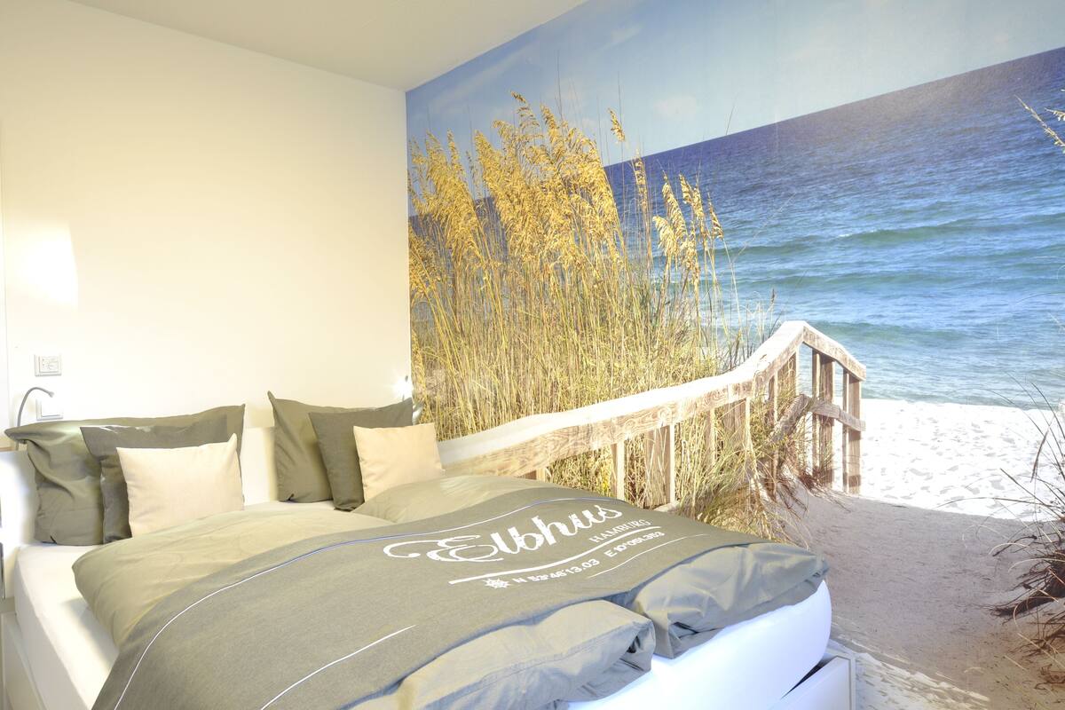 Quarto com cama, almofadas e um mural de praia na parede.