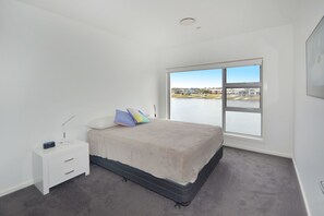 A bedroom with a large window, a bed, a bedside table, and a view of a body of water and buildings. - 4/84 The Anchorage, (Port Macquarie)