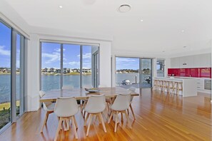 A modern kitchen with white cabinets and a red backsplash, large windows offering a view of a lake and buildings, and a dining area with a wooden table and white chairs. - 4/84 The Anchorage, (Port Macquarie)
