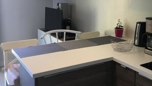 A compact kitchen with a white countertop, dark cabinets, a small dining area with a chair, a hanging light fixture, and a wall-mounted cabinet.