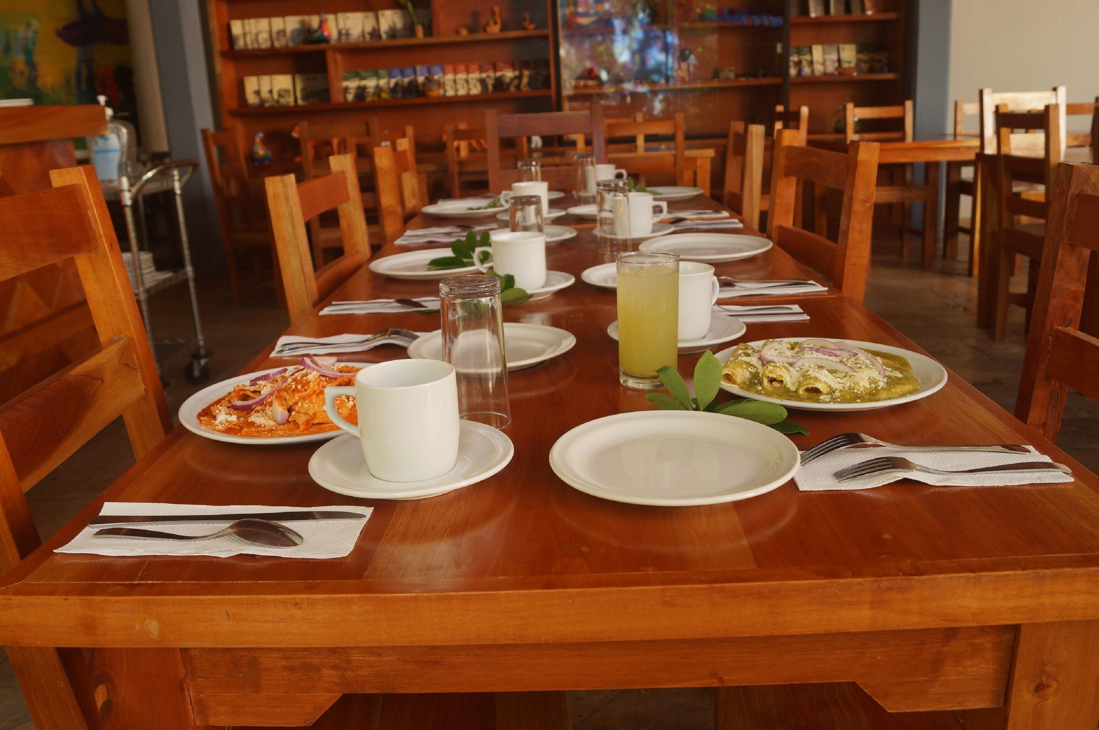 A dining table set with plates, cups, and cutlery, surrounded by wooden chairs in a restaurant with bookshelves in the background.