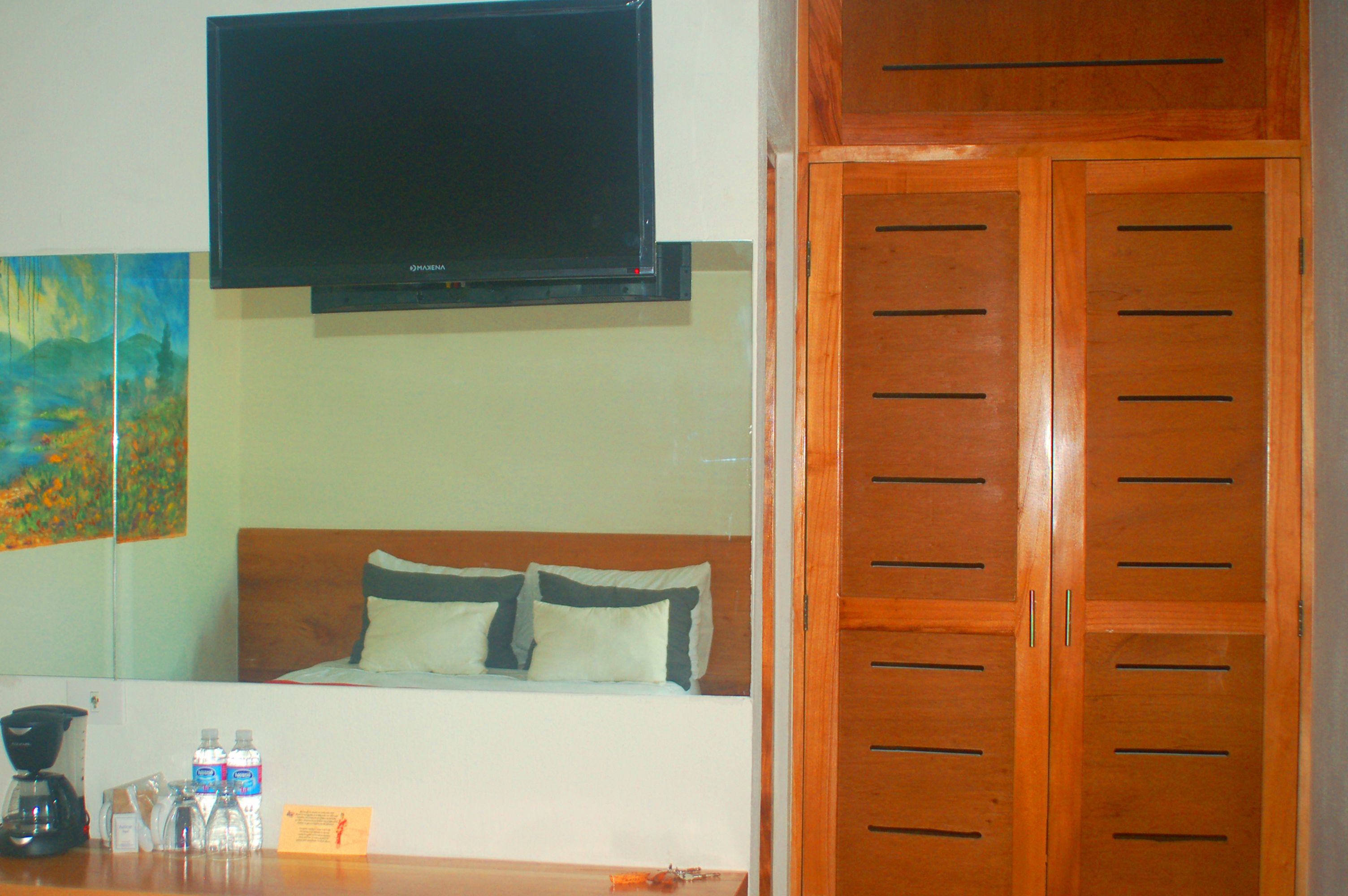 Habitación de hotel con cama, televisor montado encima, puertas de armario de madera y un cuadro en la pared.