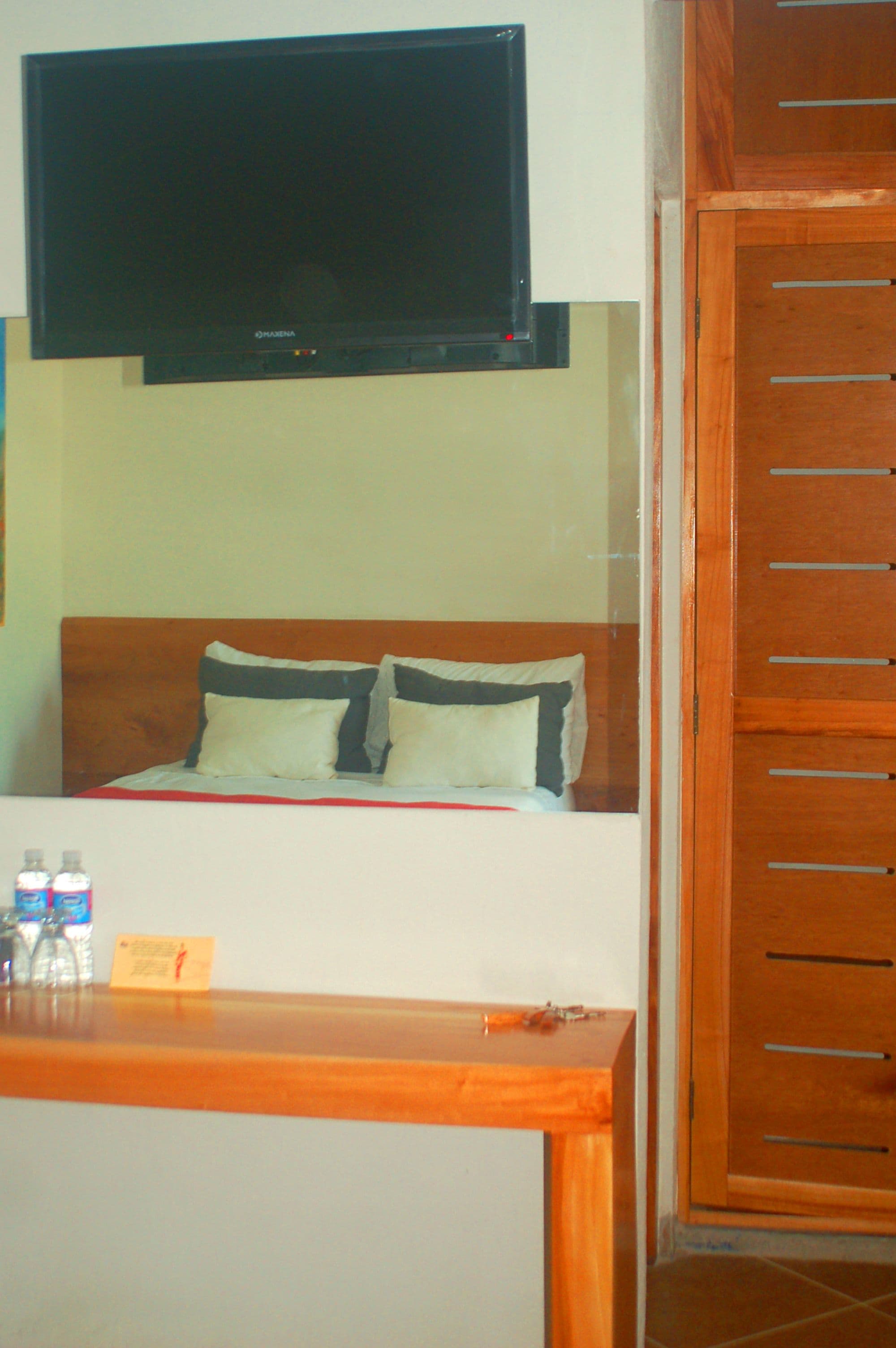 A hotel room with a bed, a TV mounted above it, a wooden cabinet, and a small table with bottles and a card.