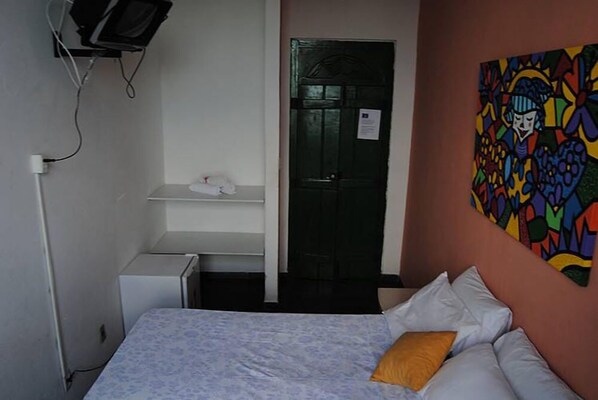 A bedroom with a bed, pillows, a small desk, a chair, a television mounted on the wall, and a door. - Pousada Solar dos Romanos (Salvador)