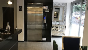 A reception area with a modern elevator, a reception desk, a decorative chair, and a view of the street outside. - Hotel Express 53 (Bogotá)