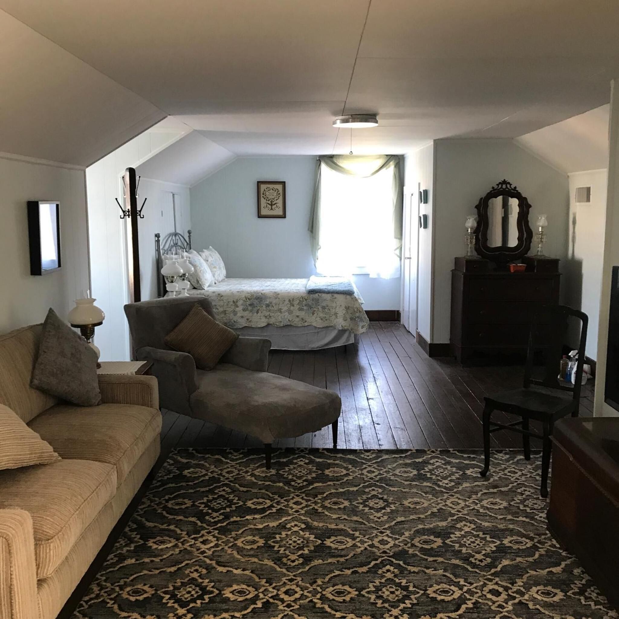 A cozy living room with a sofa, armchair, and a patterned rug. A bed with a floral bedspread is in the background, and a dresser with a mirror is to the right.