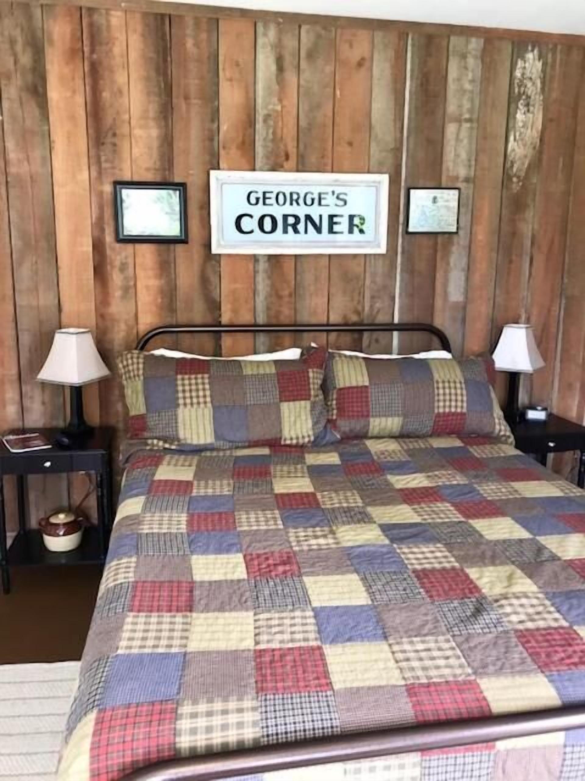 Un dormitorio con pared de madera, una cama con colcha a cuadros, dos mesitas de noche con lámparas y un cartel que dice 'GEORGE'S CORNER'.