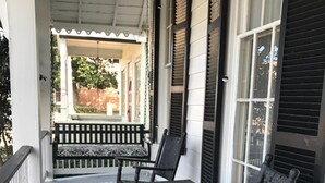 A porch with a bench and two rocking chairs, a white railing, and a view of a brick wall and trees. - Riverboat Bed & Breakfast (Natchez)