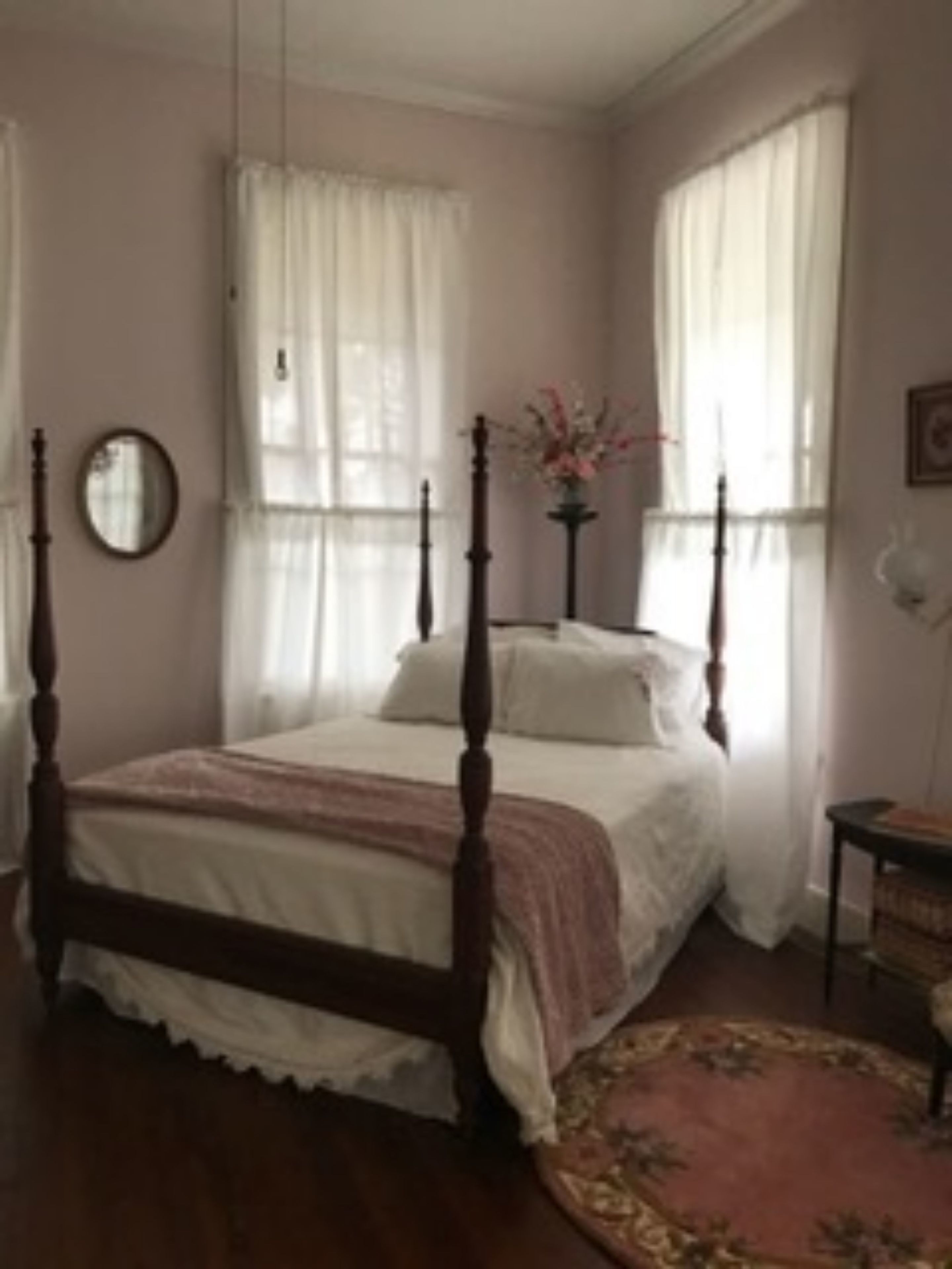 A bedroom with a four-poster bed, a bedside table, a mirror, and a window with curtains.