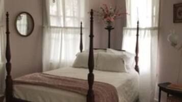A bedroom with a four-poster bed, a bedside table, a mirror, and a window with curtains.