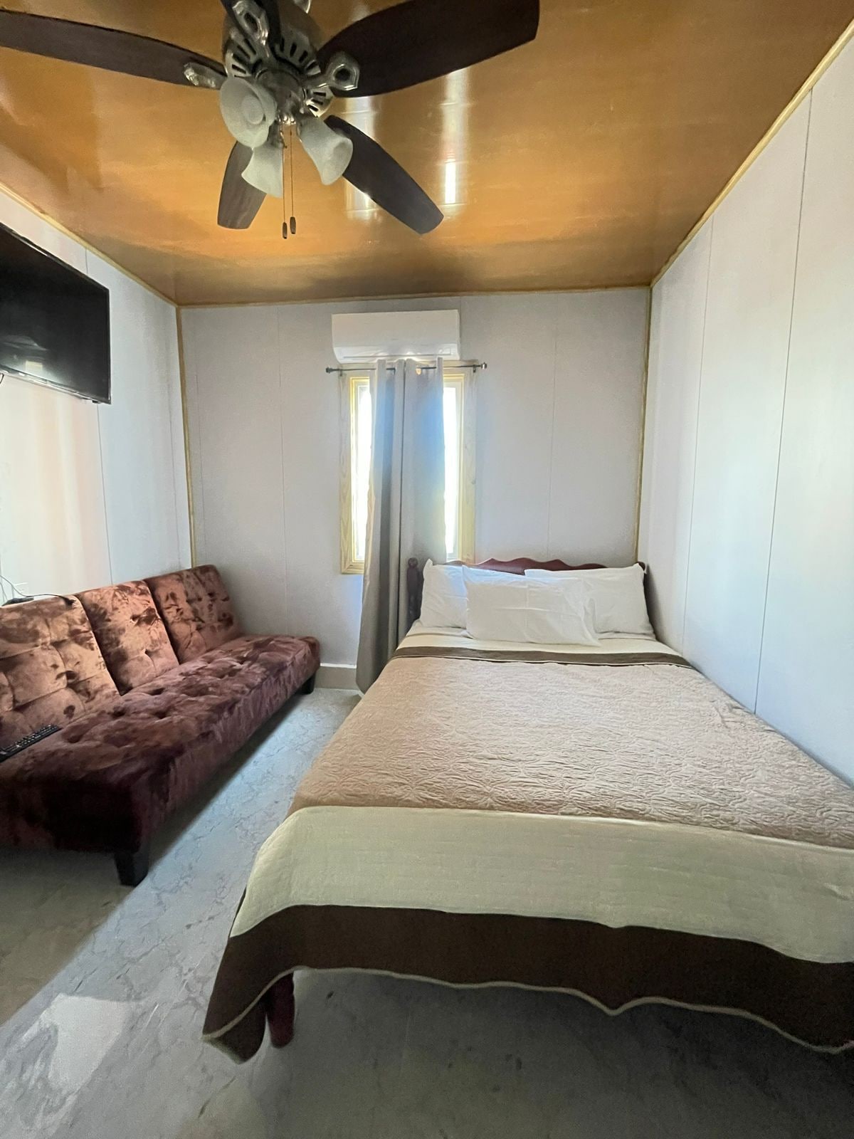 Een slaapkamer met een bed, een slaapbank, een plafondventilator en een raam met gordijnen.