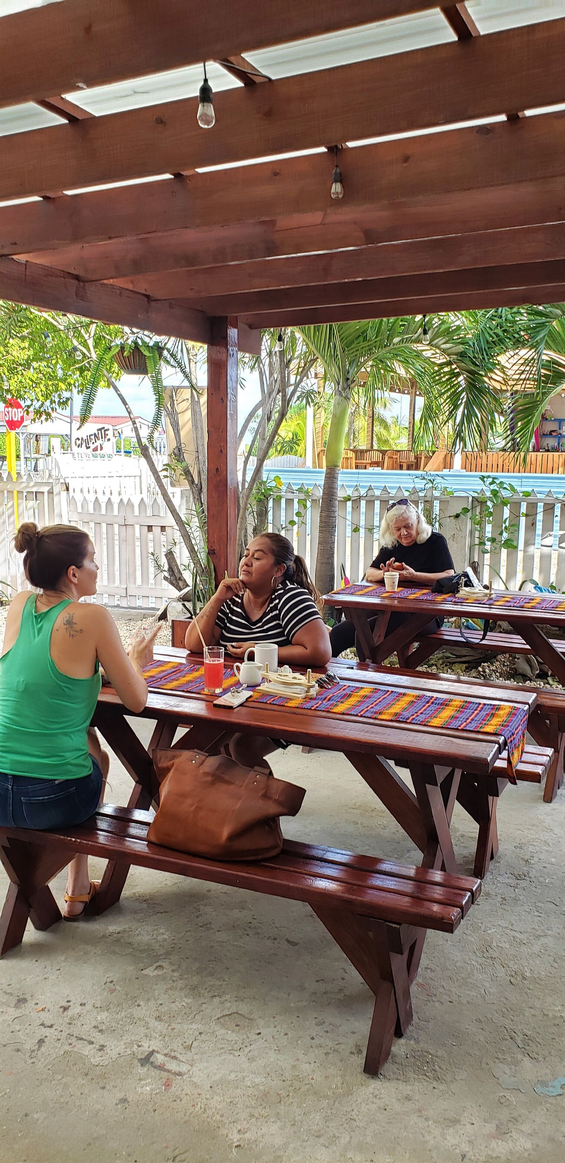 A covered outdoor seating area with wooden tables and benches, surrounded by palm trees and a white picket fence.