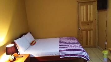 Una habitación pequeña con cama individual, puerta de madera, mesita de noche con lámpara y una colcha a rayas en la cama.