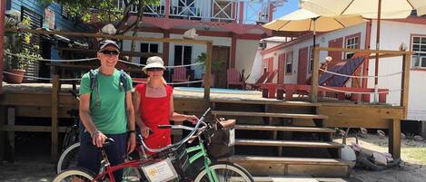 A man and a woman standing next to a bicycle with a basket, in front of a building with a pool and outdoor seating.