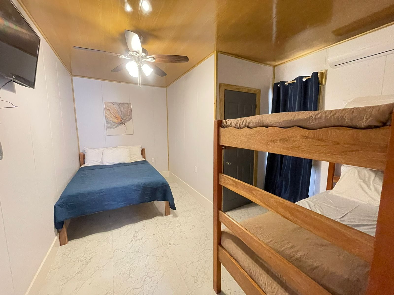 A room with a bunk bed, a single bed, a ceiling fan, a door, and a wall-mounted TV.
