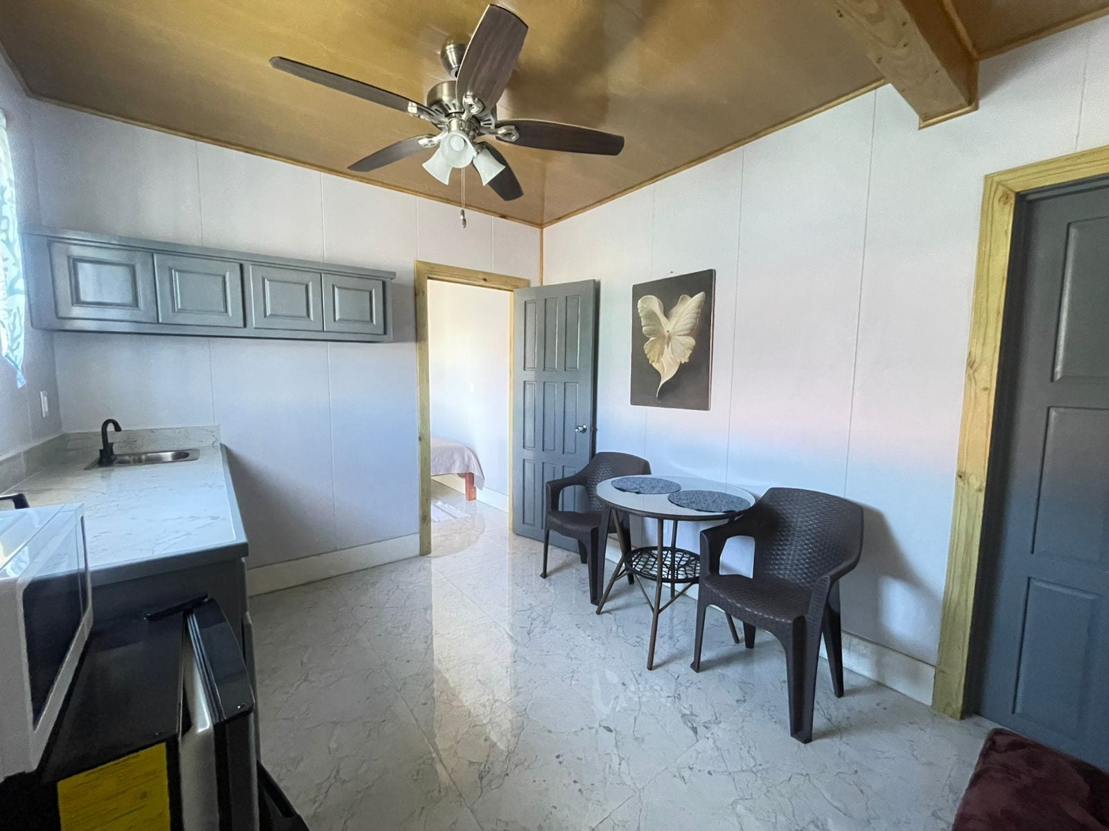 Cocina compacta con ventilador de techo, zona de comedor con dos sillas y una mesa pequeña, y una puerta que da a otra habitación.